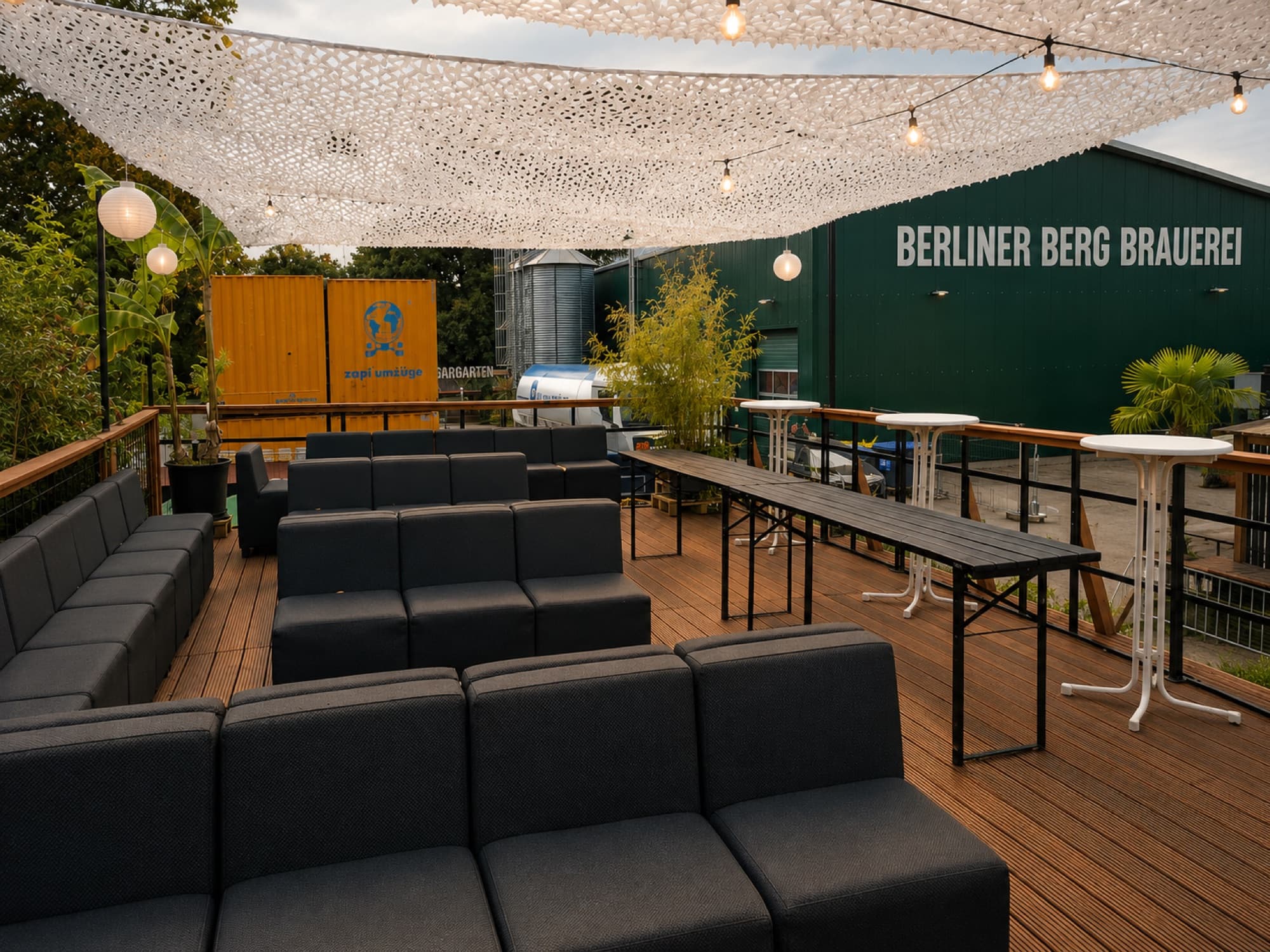 The press area at Berliner Berg Brauerei: a raised wooden deck with sofas, long tables and string lights under a white canopy, looking across to the green brewery hall.